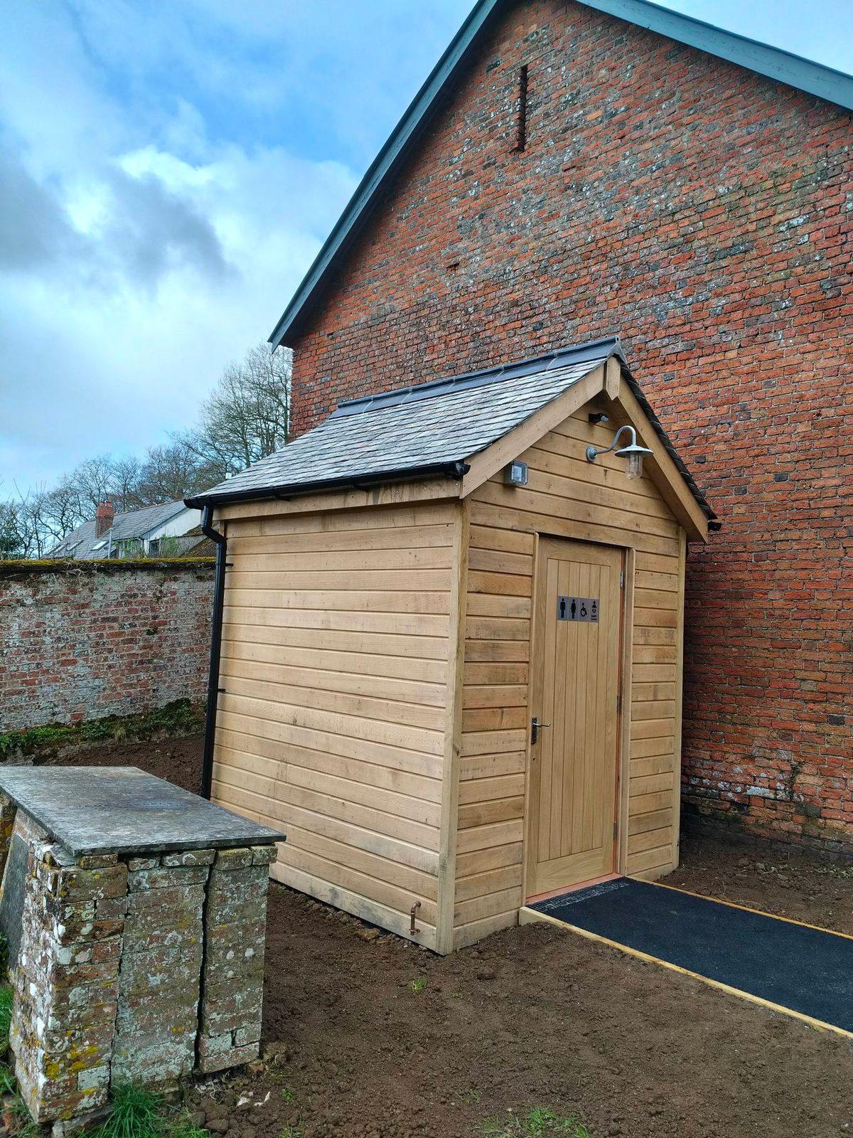 Timber-clad outbuilding with slate roof beside a brick wall